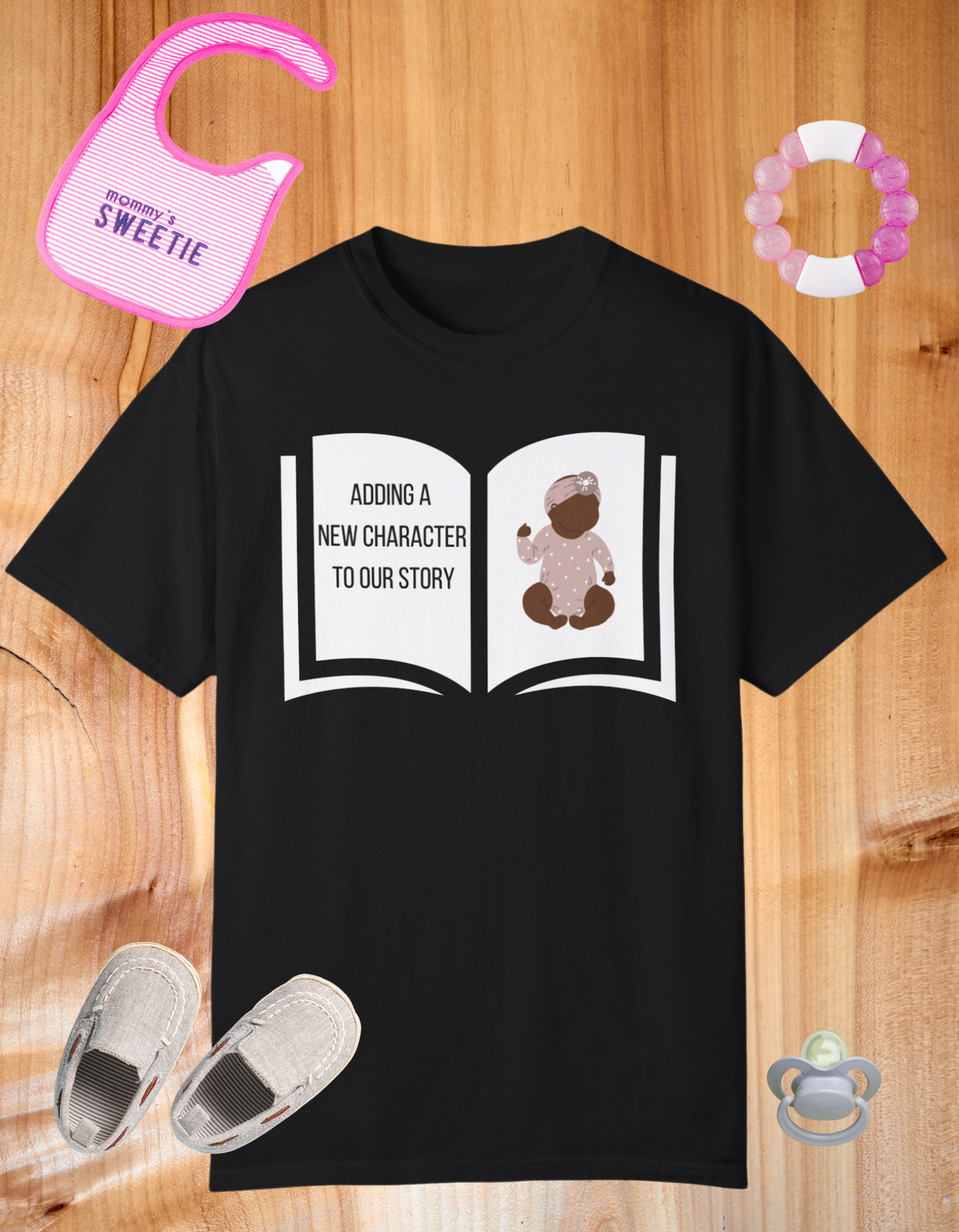 Black t-shirt featuring an open book graphic with the text "Adding a New Character to Our Story" and an image of a crib/baby booties/teddy bear, symbolizing a pregnancy announcement.