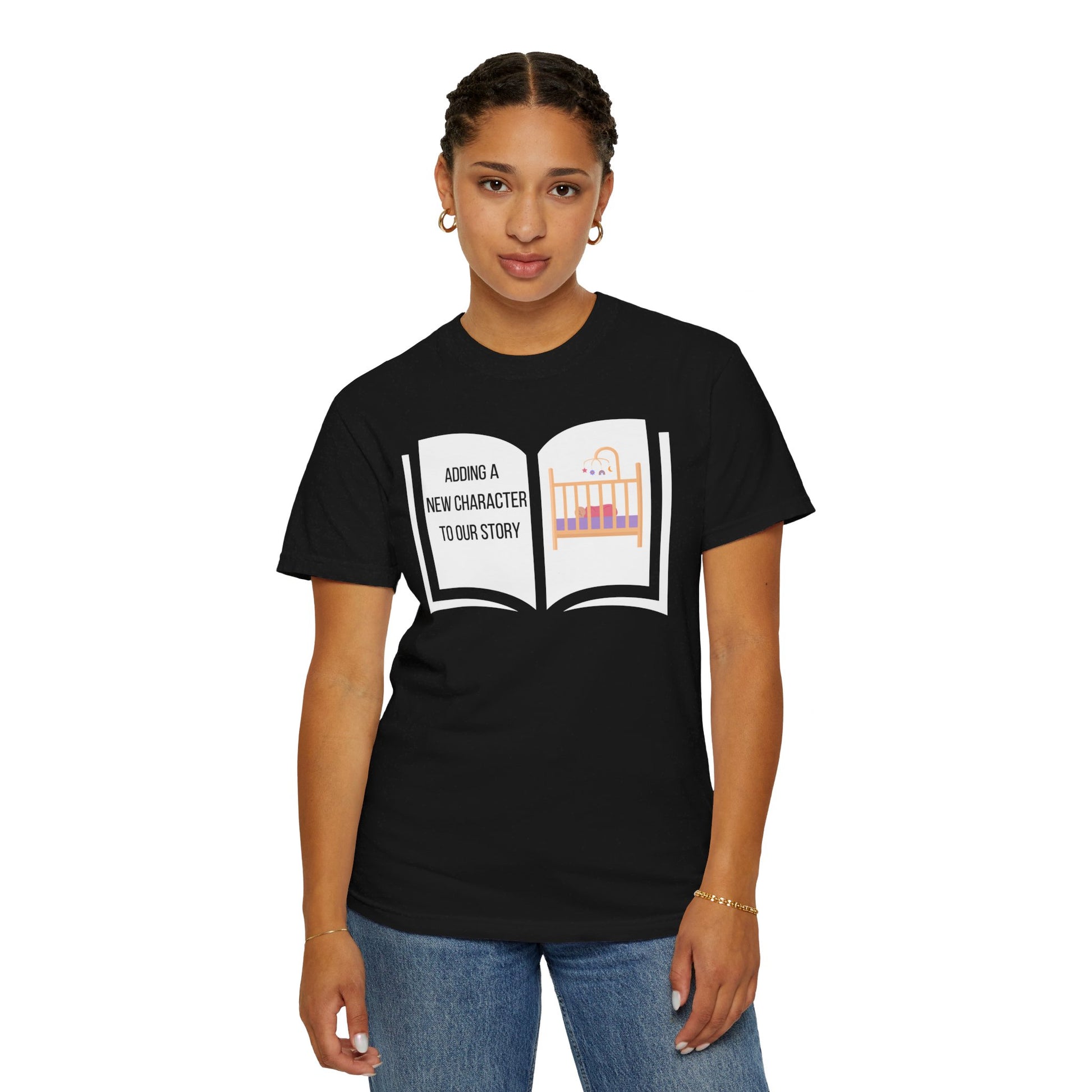 Black t-shirt featuring an open book graphic with the text "Adding a New Character to Our Story" and an image of a crib/baby booties/teddy bear, symbolizing a pregnancy announcement.