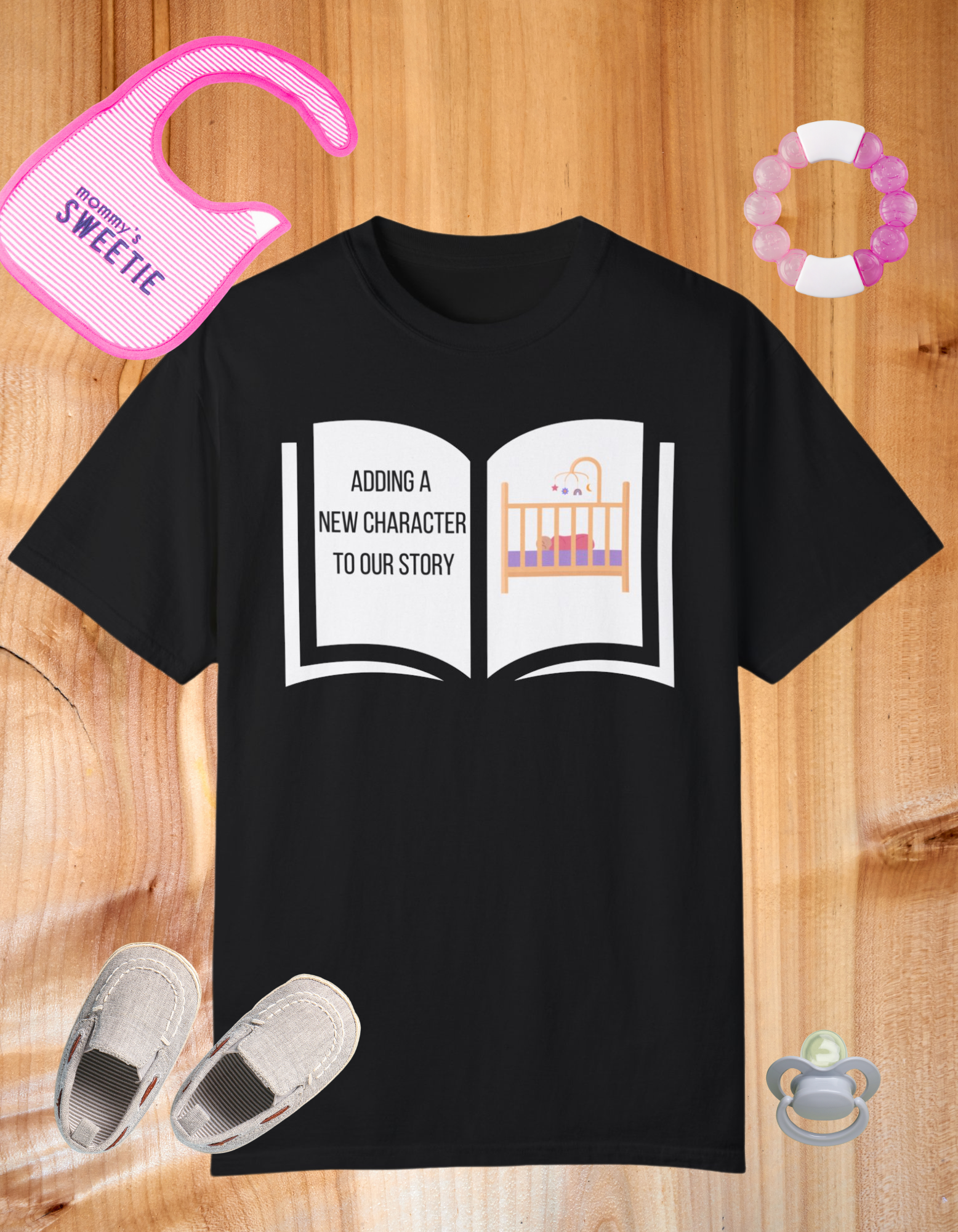 Black t-shirt featuring an open book graphic with the text "Adding a New Character to Our Story" and an image of a crib/baby booties/teddy bear, symbolizing a pregnancy announcement.