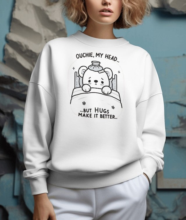 Cute white sweatshirt featuring a line-art illustration of a bear with a sore head, text above 'Ouchie, My Head,' and below 'But Hugs Make It Better.'